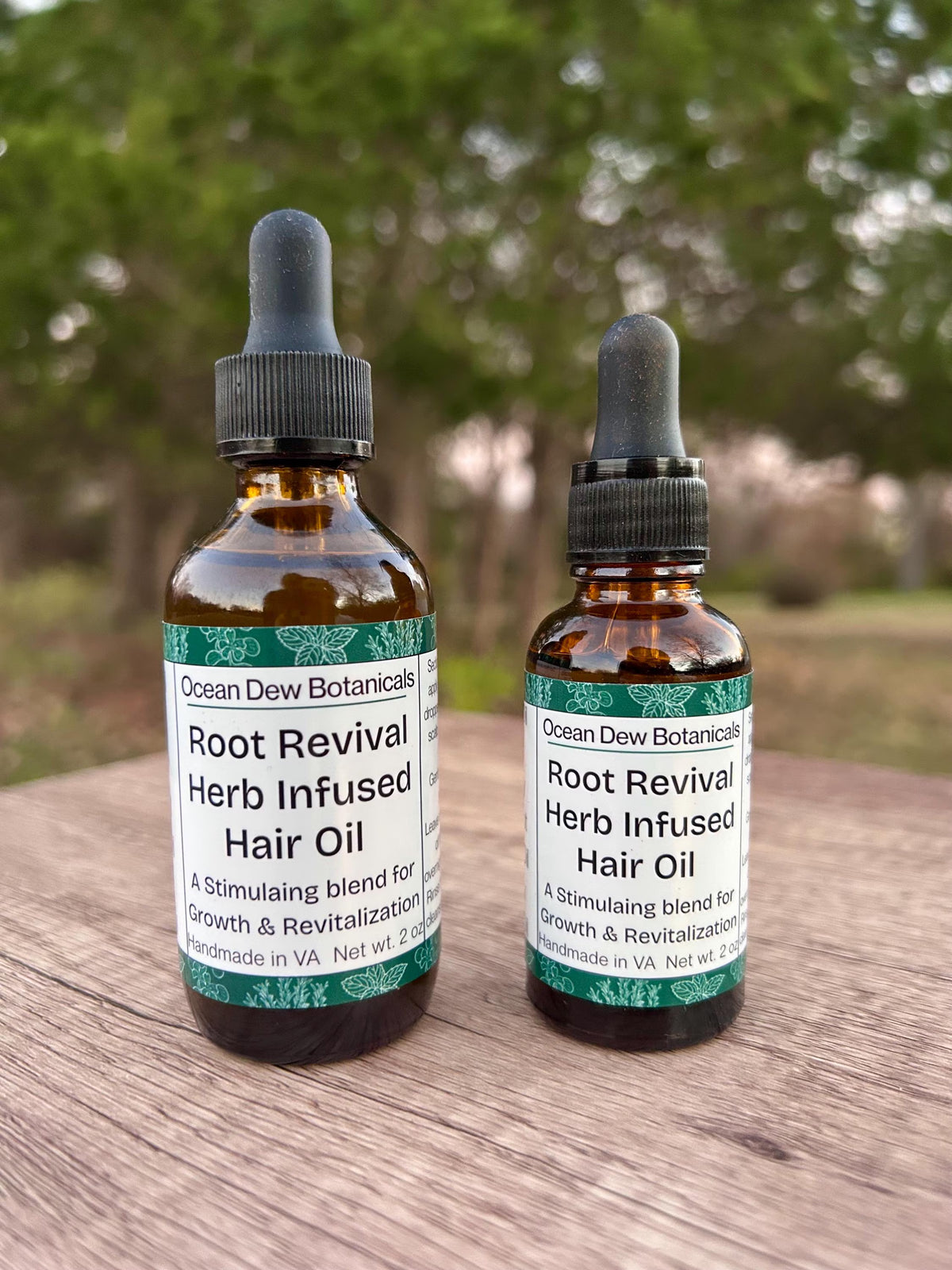 Two bottles of Ocean Dew Botanicals Root Revival Herb Infused Hair Oil on a wooden surface with a blurred natural background.